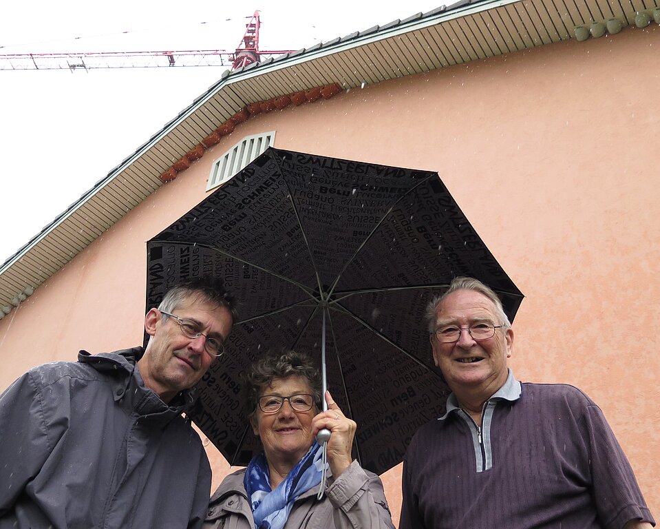 Vogelschützer Andres Beck, Susanne Hobbs, Fernand Herrmann (v.l.). Unter dem Vordach der Turnhalle haben sie Kunstnester für Mehlschwalben (links und rechts, grau) und Mauersegler (im Giebel, rötlich) montiert.Foto: Mü
